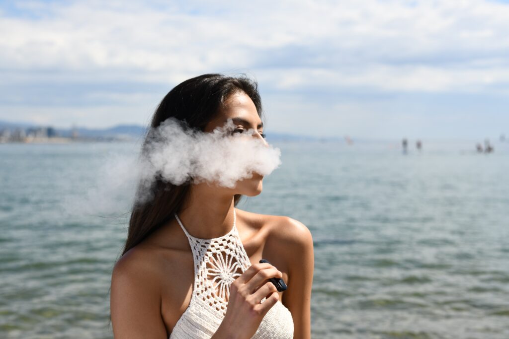 A woman enjoying a vape at the beach, with the sun setting behind her.